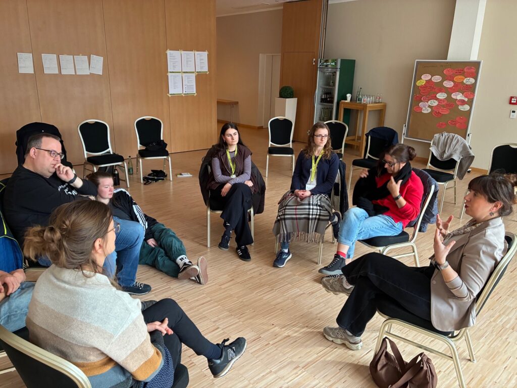 Im Kreis sitzende Kleingruppe hört einer Referentin interessiert zu.