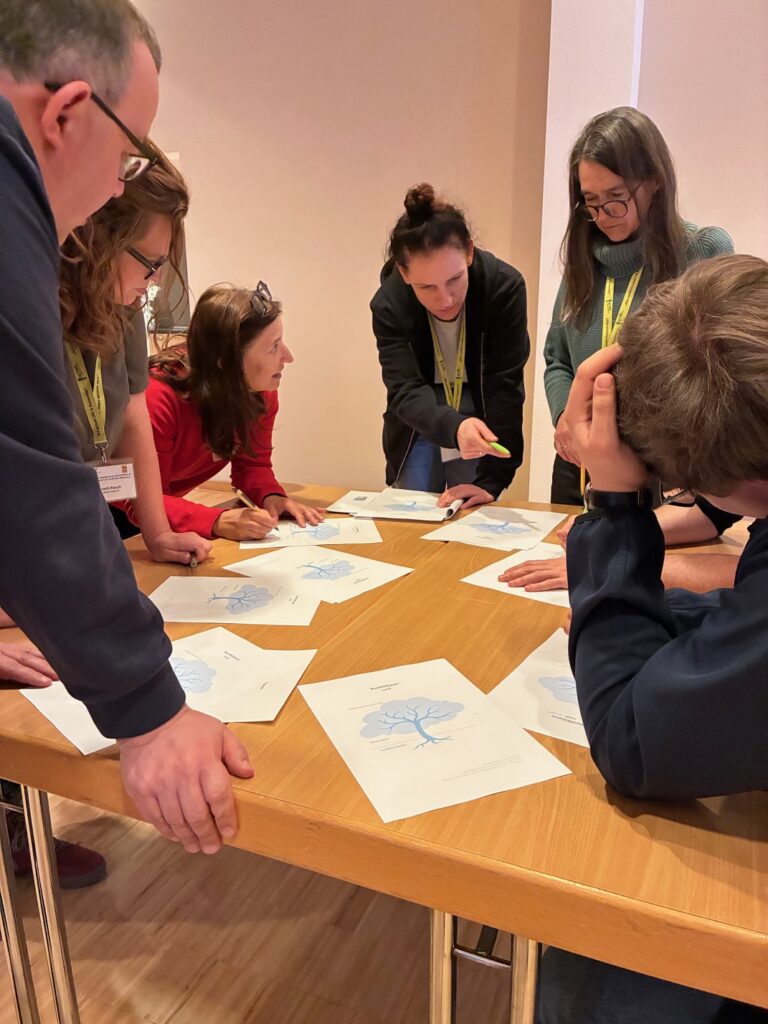 Kleingruppe steht um einen Tisch herum und arbeitet gemeinsam an ausgelegten Arbeitsblättern.